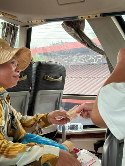 In the city of Palai, Laos, a woman wearing a straw hat and a patterned shirt is being handed money. She is seated inside a vehicle, likely a bus, with dark grey seats visible behind her. Outside the window, a tiled roof is visible, suggesting an urban or semi-urban setting. The interaction appears to be a transaction, possibly payment for a service or goods. The environment is lit by natural light, and there is a plastic water bottle on a shelf below the window.