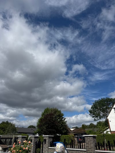 The image depicts a cloudy sky above a suburban residential area, likely within Epping Forest, United Kingdom. The foreground features a dark brick wall with a black metal fence, adorned with a cluster of peach-colored roses on the left. Beyond the fence, several houses with pitched roofs are visible, surrounded by dense green foliage and trees, one of which has a dark red canopy. A white house with red brick accents is partially visible on the right. A parked black car is seen behind the fence in the middle ground. The sky is predominantly blue with varied cloud formations, ranging from dense, dark grey clouds on the left to wispy, white cirrus clouds on the right. The overall impression is that of a daytime outdoor scene in a well-maintained residential neighborhood.