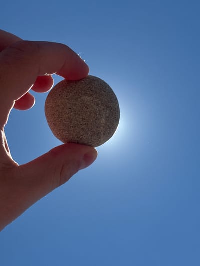 A hand is holding a smooth, gray, round stone against a bright blue sky. The sun is visible behind the stone, creating a lens flare effect. The image appears to be taken from a low angle, looking up. There are no specific geographical markers visible in the image that would indicate the location in Sudak, Ukraine.