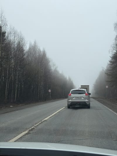 The image captures a misty landscape featuring a light-colored LADA SUV driving away from the viewer on a damp asphalt road. The road stretches into the distance, flanked by dense rows of bare deciduous trees that gradually fade into the heavy fog or atmospheric haze. The sky is uniformly light and overcast, blending almost seamlessly with the low visibility conditions.

On the rear of the SUV, a Russian license plate is clearly visible, displaying the alphanumeric code "Y278BX 18". The brand "LADA" is also visible on the tailgate. Further down the road, on the right side, a red and white circular road sign is posted. The sign depicts two cars (one red on the left, one black on the right) with a diagonal line through them, indicating a "No Overtaking" restriction. The photograph appears to be taken from inside another vehicle, with a portion of its right-side frame visible in the foreground. The overall atmosphere is dim and muted due to the dense fog, suggesting a cool day in late autumn or winter. The location is Bolgury, Russia, as indicated by the region code on the license plate.