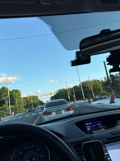 The image depicts a view from inside a vehicle, likely a silver SUV, driving through a suburban area. The driver's dashboard is partially visible, showing a speedometer indicating a speed of approximately 45-50 mph and a fuel efficiency display. Ahead, traffic is visible on a multi-lane road, with several cars including a white BMW sedan and a black SUV. Traffic lights are red at an intersection. The road is lined with trees and utility poles, and orange traffic cones suggest ongoing construction or roadwork. The sky is clear blue with scattered white clouds. A street sign visible on the right reads "KIRKHAM". This location is identified as Kirkham Drive in the city of Vaughan, Canada, specifically near the intersection with Jane Street, a major arterial road in the area.
