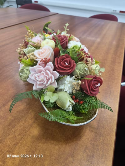 The image displays a close-up view of a decorative floral arrangement, likely composed of candles, set in a shallow, gold-colored bowl with a silver rim. The arrangement features an assortment of meticulously crafted flower-shaped objects in various colors, primarily deep red, light green, pale pink, and white. Visible wicks in the center of many flowers, such as the red roses and green buds, suggest they are candles.
Interspersed among the candle flowers are artificial green fern leaves, sprigs of small white filler flowers resembling baby's breath, and other small red and green decorative elements, adding to the bouquet's realistic appearance. One prominent candle in the foreground is a pale pink, multi-petaled flower, possibly a succulent or dahlia.
The bowl rests on a smooth, warm brown wooden surface, which appears to be a desk or table, showing subtle grain and minor wear. In the background, to the upper left, a white wall or partition is partially visible, along with a glimpse of a reddish-brown object, possibly a chair or another piece of furniture.
The photograph captures the arrangement indoors, likely during the daytime. Visible text on the bottom right of the image reads "02 юни 2026 г. 12:13," indicating the photo was taken on June 2, 2026, at 12:13 PM. The location context provided for the media file is Gara Elin Pelin, Bulgaria.