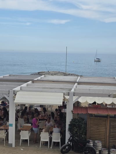 The image captures a vibrant coastal scene, likely in Sant Andreu de Llavaneres, Spain, due to the presence of the sea and coastal architecture. In the background, the sea extends to the horizon, dotted with a sailboat and a yellow buoy. A rocky breakwater divides the sea from a beachside establishment.

The foreground is dominated by an open-air restaurant or beach club, defined by a white wooden pergola structure. Underneath the pergola, numerous people are seated at tables, suggesting a meal or gathering. Most individuals are facing away from the camera, precluding detailed facial descriptions, but they appear to be a mix of ages and attire appropriate for a casual, seaside setting.

To the right, partially visible, is a building with wooden walls, possibly a bar or storage area for the establishment. Cylindrical beer kegs are stacked alongside the building, next to a modern electric bicycle. White beach umbrellas provide shade for the patrons. The scene is bathed in daylight.