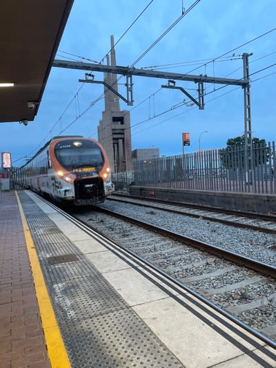 A train approaches a station platform, its orange and white exterior marked with some graffiti. Overhead electrical lines dominate the upper-left portion of the image. Adjacent to the tracks is a gray platform with a yellow safety line. A partially constructed building and fencing stand in the background behind the train. The sky is overcast.