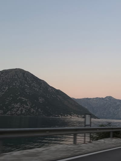 The image captures a serene coastal mountain landscape, likely at dusk or dawn given the soft pastel hues in the sky. The foreground features a road with a metal guardrail, suggesting a viewpoint from a moving vehicle. Beyond the guardrail, a tranquil body of water reflects the muted light. The background is dominated by imposing, rugged mountains covered in dark green vegetation, with some rocky outcrops visible. A small, picturesque town with white buildings is nestled along the shoreline at the base of the mountains. The overall scene evokes a sense of peace and natural beauty in a coastal region. The specific location in Kostanjica, Montenegro is characterized by its stunning bay and mountainous terrain.