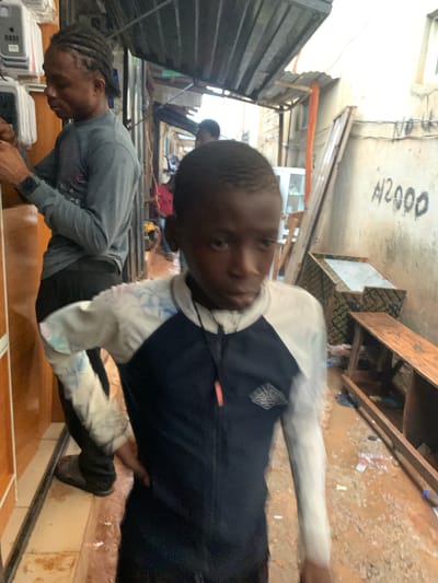 In Ado, Nigeria, a narrow outdoor market alleyway is captured. The ground is wet and muddy with scattered debris. On the left, a man with braided hair, wearing a grey long-sleeved shirt and dark pants, is attending to electrical meters mounted on a textured wall. To the right of the man, a young boy, the primary subject, is walking towards the camera, looking directly at it. He is wearing a blue and white long-sleeved shirt with a pattern on the sleeves. In the background, other people are visible, and stalls with various items and awnings suggest commercial activity. A concrete wall on the right side of the alley is marked with "N2000" in black paint. Wooden planks and shelves are visible, indicating the presence of goods or construction materials. The overall scene depicts a busy, informal marketplace environment.