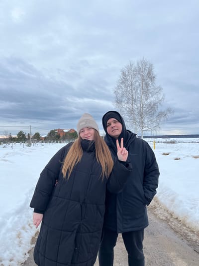 This image depicts two individuals, one female and one male, standing on a partially cleared path within a snow-covered landscape near Kalinino, Russia, under an overcast sky.
The female, positioned on the left, has long light brown hair, a light beige knit beanie, and a long black quilted puffer coat. She faces forward, exhibiting a slight smile. The male, positioned on the right and slightly behind the female, wears a black hooded jacket, a black beanie under the hood, and dark pants. He holds his right hand up, forming a "peace" sign with his index and middle fingers.
The ground is covered in white snow, with a dirt path running between the subjects and into the background, revealing patches of exposed brown earth. In the left background, a fence line, several small evergreen trees, a two-story building with a reddish roof, and utility poles with lines are visible. The right background features an expansive snow-covered field, a prominent bare birch tree with white bark, a yellow marker pole, and a distant line of dark trees. The sky is uniformly grey and overcast.