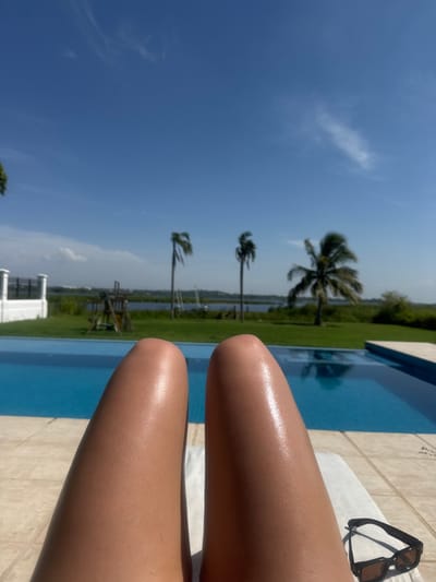 A first-person view captures the tanned, bare legs of an individual reclining by a swimming pool in Miramar, Mexico. The legs, appearing glistening, rest above a white towel in the immediate foreground, with dark-framed sunglasses positioned on the light-colored tiled deck to the lower right.
The midground features a clear blue swimming pool, likely an infinity edge, bordering a strip of green grass. Beyond the grass, a body of water is visible with several boats, including sailboats. Three distinct palm trees with varying foliage stand against a clear, bright blue sky with minimal high-altitude clouds. To the left, a white fence with pillars and a wooden playset are present on the grass. The scene is illuminated by direct sunlight, indicating daytime.