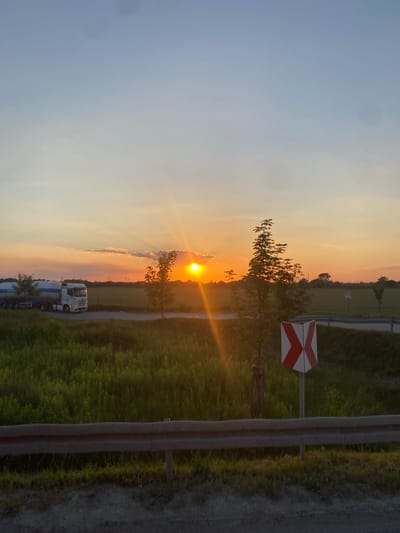 The image shows a rural road scene in Podłęże, Poland, at sunset. The sun is low in the sky, casting a warm, golden light across the landscape. A white truck with a blue trailer is parked on the side of the road, its headlights illuminating the scene. Tall green grass lines the road, and a solitary tree stands in silhouette against the setting sun. A red and white railroad crossing sign is visible to the right of the road. The sky is a gradient of soft blues and oranges, with wispy clouds catching the last rays of sunlight. The overall mood of the image is peaceful and serene, evoking a sense of calm and tranquility. There is no visible text in the image.