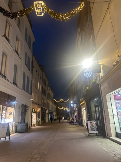 A street is adorned with festive Christmas lights. Tall buildings line both sides, and lights stretch across the street. People can be seen walking in the distance. The sky is a dark blue, and the light fixtures illuminate the scene. Shop signs are visible along the storefronts. The street appears to be paved with cobblestones or a similar material.