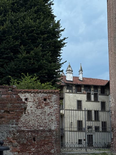 This image depicts a view of the Castello di Lagnasco in Lagnasco, Italy. In the foreground, a section of a crumbling red brick wall with a terracotta tiled cap is visible on the left. To the right of this wall is a tall, wrought-iron gate, behind which a cream-colored building with a red-tiled roof can be seen. The building has multiple windows, some with decorative arches and small painted figures above them. Two distinct white chimneys with conical tops rise from the roof. A large, dark green tree dominates the upper left portion of the image, with its foliage obscuring part of the sky. A narrow strip of green foliage is visible between the brick wall and the building. The sky is overcast with light gray clouds. A tall brick structure, likely part of the castle complex, is visible on the far right edge of the frame.