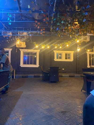 This image depicts an outdoor or semi-outdoor seating area within the city of Igbessa, Nigeria, at night. The environment features a dark-colored wall with three decorative white-framed window-like structures. Two white split-unit air conditioners are mounted high on the wall. The ceiling structure consists of exposed metal beams draped with artificial green, orange, and blue foliage, along with multiple strings of warm yellow incandescent-style light bulbs providing illumination.
The floor is a dark-colored stamped concrete with patterns resembling wooden planks and geometric tiles. Along the base of the wall, a low curb or border is painted with alternating dark and light gray or blue stripes.
One male individual with dark skin and short dark hair is visible on the left side of the frame, from the torso up. He wears a dark short-sleeved shirt over camouflage-patterned trousers and stands behind a dark, reflective counter or table, appearing to focus on an item in front of him. A small light-colored side table is partially visible to his right.
In the mid-ground, a dark-colored wicker-style table is surrounded by two matching chairs. Additional dark wicker chairs and tables are partially visible on the far right side of the frame, one of which shows a portion of a dark-skinned leg, indicating a seated person out of view. No direct interaction between individuals is discernible.