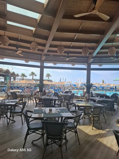 The image displays an interior view of a restaurant or dining area looking out onto a vibrant outdoor resort scene.

Indoors, the space features an aesthetically pleasing ceiling with textured, woven panels interspersed with wooden beams. Several small, light-colored ceiling fans and wicker-style pendant lights are suspended from the ceiling. The floor is composed of light-toned wooden planks, upon which sunlight casts distinct shadows. Numerous dining tables with glass tops are arranged, set with glasses and cutlery. The chairs accompanying these tables have a distinctive black and white checkered woven pattern.

Through a large glass wall that separates the indoor and outdoor spaces, a bright, sunny resort environment is visible. A large swimming pool is central to the outdoor view, surrounded by numerous blue and white lounge chairs and beach umbrellas, many of which are occupied by people relaxing. Several tall palm trees are scattered throughout the poolside area. Further in the distance, what appears to be a beach or the sea can be seen under a clear blue sky, along with more resort buildings and small Greek flags.

The scene depicts a daytime setting, likely late morning or early afternoon, characterized by bright sunshine. The overall atmosphere suggests a leisurely holiday or vacation spot.

The text "Galaxy A55 5G" is visible in the bottom right corner of the image.