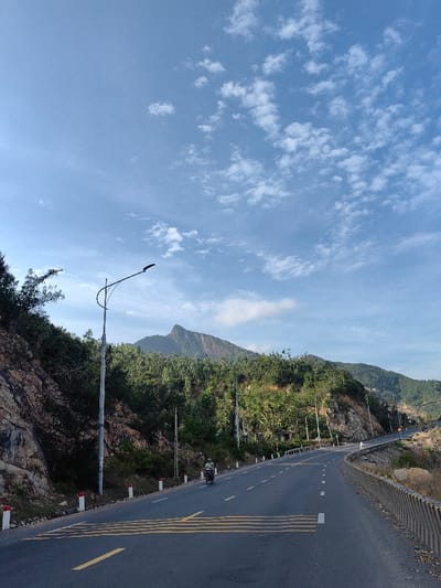 A paved two-lane road, marked with solid yellow and dashed white lines, including a series of thick yellow horizontal lines in the foreground, curves right and ascends through a mountainous region of Tuy Phong 4, Vietnam. White and red bollards line the left shoulder, while a concrete barrier is visible on the right. Rocky terrain and dense green foliage cover the hillsides, with a prominent, jagged mountain peak rising in the background. Multiple streetlights with dark, curved fixtures are positioned along the roadside. One individual, wearing a light-colored shirt and a helmet, operates a white scooter or motorcycle, traveling away from the viewer in the left lane. The sky is clear blue with scattered white clouds.