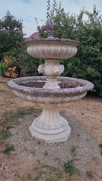 The image depicts a two-tiered, ornate stone fountain situated outdoors. The fountain is a creamy beige color, with a weathered appearance showing some darkening and staining on the surfaces. The upper basin is smaller and more intricately decorated, topped with a decorative finial resembling a fleur-de-lis. The lower basin is wider and deeper, with decorative carvings around its rim and several carved lion heads projecting from the central support column. The lower basin contains some dark-colored ground cover, possibly mulch or soil, and a cluster of dark, reddish-brown, cone-shaped objects, possibly dried pinecones or seed pods, in the foreground. The fountain stands on a base surrounded by gravel and dry, brown grass. In the background, lush green trees and bushes create a dense natural setting. A portion of a building with a reddish-pink roof is visible through the foliage. The sky above is overcast with soft clouds. The overall impression is of a garden feature in a rural or suburban environment. The specific location in Macé, France is not discernible from the image itself.