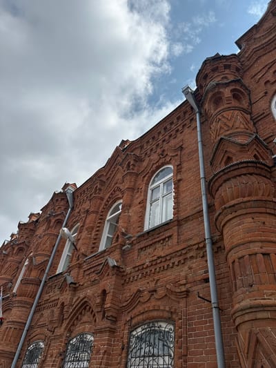 The image shows the exterior of a red brick building with ornate architectural details, viewed from a low angle looking upwards towards a cloudy sky. The building features arched windows with white frames and decorative wrought-iron grates on the lower level. The brickwork is detailed with various patterns and motifs, including what appear to be small turrets or decorative towers. A grey downspout runs vertically along the side of the building, and a lamppost is attached to it. The sky is partly cloudy with patches of blue visible. The location is identified as Nogornyi, Russia. There are no people or actions visible in the image.
