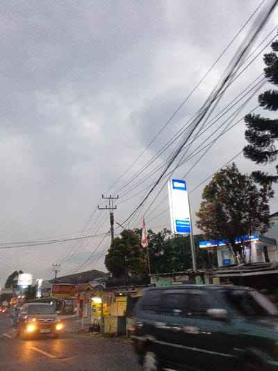 This image depicts a street scene in Phước Hòa, Vietnam, under an overcast grey sky, indicative of low light conditions such as dusk or dawn, with artificial illumination active. A paved road occupies the lower portion of the frame, displaying a yellow lane marking. On the right side of the foreground, a dark-colored minivan or SUV is in motion, blurred due to its movement from right to left. In the mid-ground on the left, a dark-colored car with illuminated headlights faces the camera, appearing stationary or moving slowly.
Overhead, numerous black electrical wires crisscross the frame, supported by multiple utility poles. Along the right side of the road, a prominent, tall, rectangular sign is illuminated with blue and white sections. Behind this sign, a building with similar blue-themed illuminated signage, possibly a convenience store or ATM, is visible. Further down the street on the left, various commercial establishments line the road, displaying illuminated signs, some partially legible, including "KONTER HP." A red and white flag is mounted on a pole near one of these buildings. Green trees are interspersed among the buildings and alongside the road, contributing to an active urban or semi-urban environment with vehicular traffic and commercial activity.