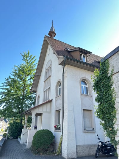 This is a vertical, eye-level shot of a white stucco building with a brown tiled roof, featuring a small, pointed spire. Ivy grows along the top of the building and down one side. There are several windows visible, some with arched tops, and one with a decorative geometric surround. A black motorcycle is parked on the paved area next to the building. A large green tree with lush foliage is to the left of the building, and a manicured green bush is to the right. The sky is a clear, bright blue, suggesting a sunny day. In the distance, other buildings and trees are visible, indicating a residential or urban setting. The overall impression is of a well-maintained property on a pleasant day.
