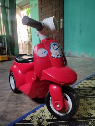 A red toy scooter sits on a patterned mat in front of a teal-colored wall and a wooden door. The scooter has a black seat, white wheels with black tires, and a white headlight. A black handlebar extends from the front. The background shows a glimpse of a window with green curtains and outdoor greenery, suggesting a residential setting. The location is identified as Puloblang, Indonesia.