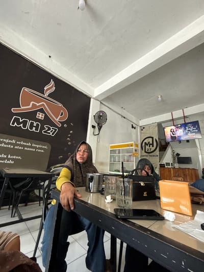 In Meunasahmanyet, Indonesia, an indoor scene captures two women seated at a table within what appears to be a cafe or eatery. The woman on the left, wearing a yellow long-sleeved shirt and a patterned hijab, is looking upwards and to the left with a stern expression. Her right arm rests on the table, obscuring part of a black, wooden edge. To her right, on the table, are two clear glasses filled with liquid, one with a black straw, a stainless steel cup, and a black handbag with a gold clasp. In the foreground, a dark-colored smartphone is placed on the table, next to a crumpled white tissue. To the right of the smartphone is an orange object with a folded corner, possibly a wallet or a book.

The woman on the right is wearing a dark green hijab and is looking downwards, partially obscured by the black handbag. In the background, a large dark banner with a stylized coffee cup logo and the text "MH 27" is visible on the wall. To the right of the banner, a fan is mounted on the wall. Further to the right, a television screen displays a program featuring two individuals. Another banner with a circular logo and the text "MH 27 COFFEE PREMIUM" hangs on the wall. A white shelf unit with various containers is also visible. Wooden paneling forms the counter area in the far background. The floor is tiled with light-colored squares.
