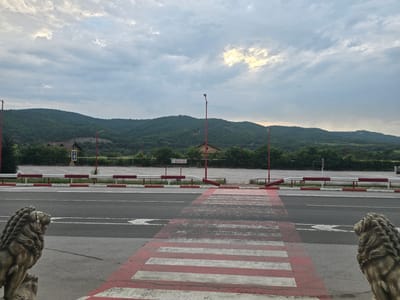 The image depicts an outdoor scene with a focus on a road and a parking lot, set against a backdrop of rolling, tree-covered hills. In the foreground, a pedestrian crossing with red and white stripes is visible, leading across a grey asphalt road. On either side of the crossing, stone lion statues are placed. The road has lane markings. Beyond the road, a white barrier with red accents separates it from a large, empty asphalt parking lot. Several red-topped lampposts are positioned throughout the scene, with one prominently in the middle ground. In the distance, a few buildings and dense green foliage are visible at the base of the hills. The sky is overcast with patches of sunlight breaking through the clouds, suggesting a late afternoon or early morning setting. The image appears to be taken in Zlatna Panega, Bulgaria, given the context provided. There are no people or animals visible in the frame.