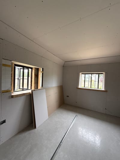 The image depicts the interior of a room under construction, located in Skipsea, United Kingdom. The room is unfurnished and features walls and a sloped ceiling largely covered with newly installed, unfinished gypsum plasterboard panels secured with visible screws. Seams between panels are present on the ceiling and walls.
Two rectangular windows are visible. The window on the left is partially framed with rough timber and shows some green mesh behind an unboarded section of the wall. The window on the right is fully framed with a light-colored wooden sill. Both windows have dark frames and offer a view of an outdoor area with greenery and fencing. A double electrical socket is installed on the wall below the right window.
Leaning against the wall below the left window are two vertically positioned plasterboard sheets: one white and one brown/tan. The floor is a smooth, light grey surface, appearing to be concrete or screed. Two long, silver-colored perforated metal channels, likely used for framing, lie parallel on the floor in the foreground. No individuals are present in the scene.