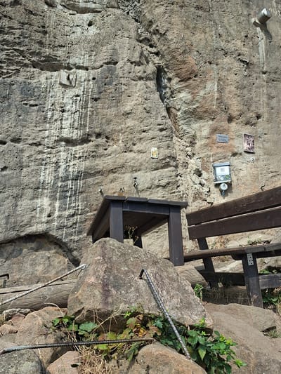 The image depicts a rocky cliff face in Děčín, Czechia, featuring a small wooden table and bench. A metal rope runs along the rocks in the foreground, anchored to a large boulder. The cliff face has markings and textures indicative of erosion and weathering. Mounted on the rock are several items: a square metal fixture, a small rectangular plaque with text, a framed image, a metal box with a QR code and a lock, and a second framed image. There are no people visible in the scene. The vegetation is sparse, consisting of grasses and small leafy plants growing around the base of the rocks. The environment appears to be outdoors, likely a natural or semi-natural recreational area.