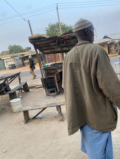 An older man wearing a kufi hat and green jacket stands beside a weathered wooden structure. The street scene includes power lines, a stall, and a building with "P.O.S. 1.15" visible. A white bucket sits nearby. Another person wearing a similar hat walks in the distance on the road. The overall atmosphere suggests a rural or less developed area.