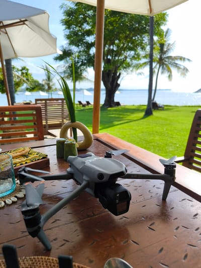The image shows a DJI Air 3S drone sitting on a dark wooden table. The drone is gray, with a black camera mounted below its body. The table is set outdoors, under a white umbrella, with a partial view of a green lawn, palm trees, and the ocean in the background. There are other objects on the table including a decorative glass, a dish with snacks, and other decorative items. The background suggests a tropical resort or beachfront property. The location is Sigar Penjalin, Indonesia. There are no people visible in the immediate foreground.