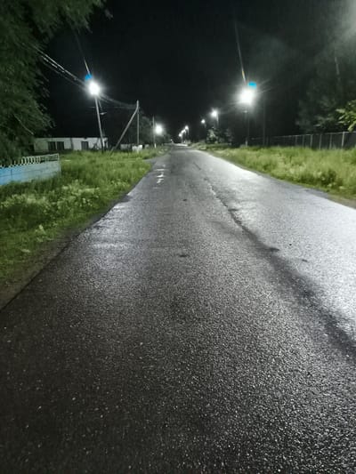 A wet asphalt road is illuminated by streetlights at night. The road is bordered by a grassy area and trees on the left and a fence on the right. In the distance, several streetlights are visible along the road, indicating it is in a populated area. The overall impression is a quiet, slightly eerie scene, possibly after rain. The location is the city of Arkalyk, Kazakhstan.