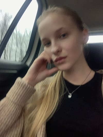 A young, fair-skinned female is depicted from the chest up, seated inside a vehicle. She has long, blonde hair, with a portion styled over her left shoulder. Her right hand is raised, with fingers resting against her chin and jawline. She is wearing a dark top, possibly black, underneath a light beige knitted cardigan with a visible ribbed texture on the sleeves. A silver-colored chain necklace with a small, rounded, light-colored pendant is visible on her chest. Her gaze is directed forward. Through the car window to her left, bare deciduous trees with thin trunks are visible against an overcast sky, suggesting a non-foliage season. The scene is located in Oliivka, Ukraine.