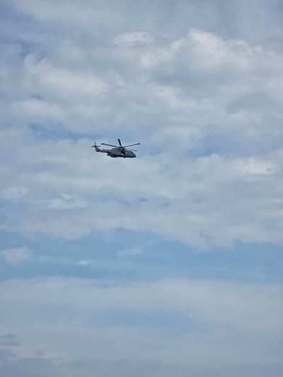 A grey military helicopter is captured in flight against a cloudy sky. The aircraft, resembling a Westland Merlin, features a multi-bladed main rotor and tail rotor, and appears to have a side door or window open. It is positioned in the upper-middle section of the frame, moving leftwards. The sky displays a light blue base with numerous scattered cumulus clouds ranging from white to light grey. The lower edge of the image shows an indistinct, darker horizontal band. The location is the city of Unknown, Sovereign Base Areas of Akrotiri and Dhekelia.