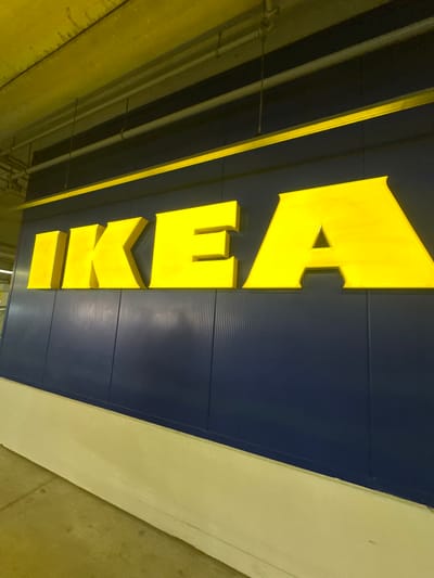 The media file shows a close-up, angled shot of the large, yellow, three-dimensional IKEA logo on a dark blue, corrugated metal facade. The letters spell out "IKEA" vertically. The lighting suggests an indoor, likely commercial, setting, possibly a parking garage or an exterior wall of a building, given the concrete structures and overhead pipes visible in the yellow-tinted areas. The perspective is from below, looking upwards at the sign. The scene is static, with no activity or people visible. The overall impression is a clear, well-lit view of the iconic brand signage. The yellow color of the text stands out vividly against the dark blue background. There are no indications of time of day or weather conditions, as the environment appears to be controlled and indoors or under cover. The location context provided is Alfafar, Spain, which is consistent with the presence of an IKEA store.