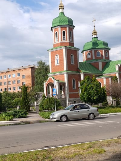 The image captures a street view in the city of Khorol, Ukraine, featuring a prominent brick church with green domes and a tall bell tower. In the foreground, a silver sedan is driving on an asphalt road across a pedestrian crossing. To the left of the church, a multi-story brick apartment building is visible, surrounded by lush green trees and bushes. The sky is partly cloudy, suggesting it is daytime. The scene is calm and depicts a typical urban landscape.
