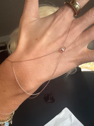 A close-up shot features a person's left hand and wrist, adorned with jewelry. On the ring finger, a thick, gold-colored band is visible. A delicate, rose-gold colored hand chain connects this ring to the wrist, spanning across the back of the hand. The centerpiece of this hand chain is a small, ornate charm shaped like a Hamsa hand, detailed with an "evil eye" design in its palm. Around the wrist, a multi-strand bracelet is partially visible, also featuring a blue "evil eye" bead among its chains.
The hand is positioned against a dark, out-of-focus background, which appears to be an indoor setting like a car interior or a table. In the upper left background, some blurred lighter areas suggest an outdoor view through a window. A piece of white paper with faint grid lines is visible in the upper right corner of the frame. Long, light-colored hair, likely belonging to the person wearing the jewelry, is visible and blurred in the lower left and center background. The skin of the hand shows natural texture and creases. The overall impression is a casual display of personal accessories, likely in Strömstad, Sweden. No text is visible in the image.