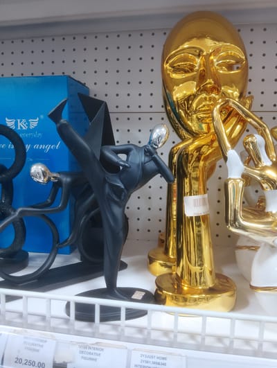 The image depicts a retail display shelf at a store in Aketu-Oja, Nigeria. The shelf holds several decorative figurines. Prominently featured are two large, shiny gold decorative pieces: one a stylized human face and the other a set of figures in an abstract pose. In front of these, and to the left, is a black matte figurine of a person in a dynamic kicking pose, appearing to be an athlete. Behind the black figurine and to its left is a portion of a blue box with white text and a logo. The objects are arranged on a white grid shelf. Pricing labels are visible on the lower part of the image, indicating product codes and prices. The background consists of a white pegboard wall with evenly spaced holes.