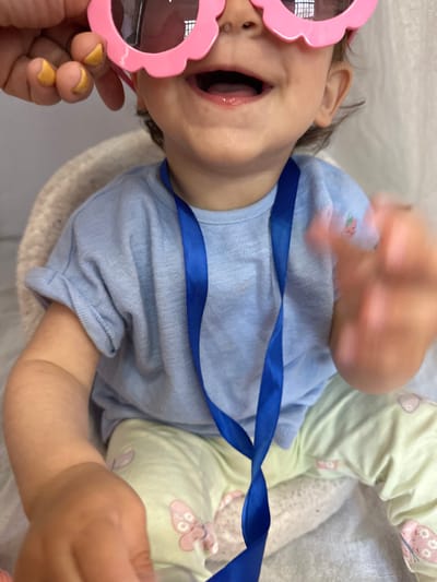 A close-up shot shows a baby wearing large pink heart-shaped sunglasses and a light blue t-shirt. A blue ribbon hangs from the t-shirt. The baby is wearing pale green pants with a butterfly pattern. The baby's mouth is open, and their hands are visible. The background is a soft white. The image does not provide any information about the location of Prestbury, United Kingdom.