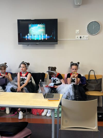 The image depicts an indoor waiting area, likely backstage or a common room, within a facility in Ventspils, Latvia. Four young girls, appearing to be pre-teen, are seated on a dark upholstered bench against a plain light beige wall.

All girls wear matching black and white Dalmatian-themed costumes. These consist of white bodices featuring large black paw print designs, layered black and white tulle skirts, and red neckerchiefs with white spots. Their hair is styled with black and white spotted headbands that resemble dog ears. Black knee pads are visible on at least two of the girls.

The girl on the far left, partially visible, is looking down at a purple Nintendo Switch Lite device. The girl to her right, in the center-left, has prominent red lipstick and looks forward with a neutral, pensive expression, not currently interacting with a device. The third girl, in the center-right, is looking down at a red Nintendo Switch Lite. The fourth girl, on the far right, is looking down at a yellow smartphone.

In the foreground, a light-colored wooden table spans the width of the frame. On the table, a silver laptop computer is open. Below the table, a red duffel bag with the number "75" printed in white is visible, along with various pairs of white sneakers and other bags. A black cushion rests on a pink suitcase in the very bottom foreground.

Mounted on the wall above the bench is a black Sony Bravia flat-screen television displaying a stage performance. The screen shows approximately seven figures in white costumes on a brightly lit stage against a light blue backdrop depicting clouds. To the right of the television, a circular white volume control dial and a white wall-mounted strip containing multiple European-style electrical sockets and a network port are visible. Further to the right on the wall, a large, round wall clock with a grey metallic frame and a light blue face shows the time as approximately 10:45. A white speaker is mounted high on the wall above these fixtures.

To the far right, a dark brown designer-style tote bag rests on a light brown chair. The floor of the room is segmented into dark brown and dark grey sections.