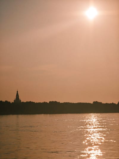 This media file depicts a serene landscape scene with a body of water in the foreground and a silhouetted tree line and a building in the background. The sun is bright and high in the sky, casting a warm, golden glow over the entire scene. The water reflects the sunlight, creating a shimmering effect.
The subjects in the scene are the natural elements: the water, the trees, and the sky. There are no people visible. The building appears to be a church with a tall spire and a cross on top, silhouetted against the sky. The tree line forms a dark band across the middle ground, suggesting a forest or dense vegetation.
The activity taking place is simply the passive observation of a natural landscape. The time of day is clearly daytime, given the bright sun. The weather appears clear and sunny. The location cues, such as the presence of a church and a dense tree line, along with the provided location context of Lesosibirsk, Russia, suggest a possibly rural or semi-rural setting in that region.
Notable details include the intense brightness of the sun, which creates a lens flare effect at the top of the image. The water's surface is textured with ripples, and the sunlight creates a dazzling reflection across it. The overall mood conveyed by the image is one of tranquility and natural beauty, enhanced by the warm, atmospheric lighting. There is no visible text in the image.