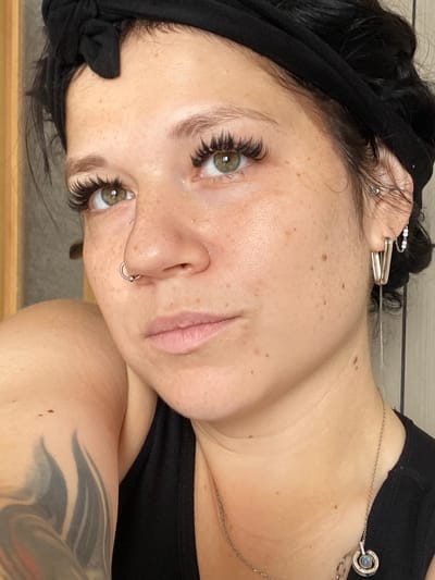 The image is a close-up portrait of a woman with freckles and green eyes, looking slightly downwards and to her left. She has on false eyelashes, a black headband tied at the top of her head, and a nose ring. She is wearing a black tank top, and a silver necklace with a pendant. Her left arm, partially visible, has a large tattoo. The background is out of focus and appears to be an indoor setting with light-colored vertical blinds on the right. The lighting suggests it is daytime. The image is cropped to focus on the woman's face and upper body. There is no information in the image to suggest its location in the city of Ostrovki, Russia.