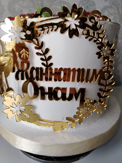 A white frosted cake is decorated with a gold floral wreath around golden Cyrillic lettering. The cake sits on a white platform with a gold ribbon around the base. The lettering is horizontally centered in the wreath. The frosting is smooth, and the gold decorations have a mirror-like sheen, reflecting the surrounding environment. The image captures a celebratory or commemorative occasion, likely a birthday.
