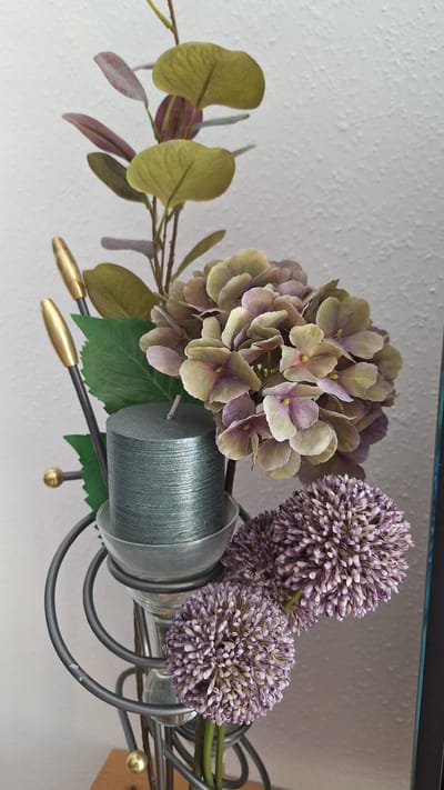 The image shows a close-up of an artificial floral arrangement, likely a home decor item, placed indoors against a textured white wall. The arrangement features a prominent cluster of muted purple and green hydrangea-like flowers. To the left of the hydrangeas, a cylindrical teal candle with a metallic sheen stands in a clear glass holder. The candle is part of a dark gray metal stand with spiraling elements and decorative gold finials at the top. In front and to the right of the candle, there are two spherical, purple ornamental onion-like flowers. Behind the main flowers, there are some artificial green and burgundy leaves and stems. The overall setting appears to be a domestic environment, possibly a living room or dining area, in Schwaikheim, Germany. No people are present in the image.