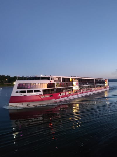 A wide, long river cruise ship, painted predominantly white and bright pink, moves from left to right across the frame, creating wakes on the dark water. The ship has multiple decks with rows of windows, many of which are illuminated with warm, golden light, suggesting occupancy. A circular logo with red and white elements is visible on the side of the ship, near the front. The sky above is a deep, clear blue, transitioning to a lighter hue near the horizon, indicating either dawn or dusk. The shoreline to the left features a dark line of trees, with some lights visible among them, hinting at a settlement. The reflection of the ship's lights shimmers on the water's surface, adding to the atmospheric depiction of the scene. The image appears to capture the vessel navigating a body of water near the city of Il'inskoe (Lomovskaya S/A), Russia.