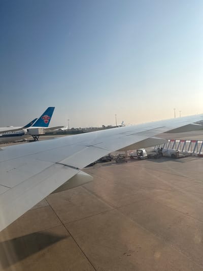 The image displays an aerial view from an aircraft window on an airport tarmac at Charles de Gaulle Airport (CDG) in Tremblay-en-France, France, under clear daylight conditions.
The foreground is dominated by the leading edge and upper surface of a white aircraft wing, which extends from the lower left to the upper right of the frame, partially obscuring the tarmac below. A distinct shadow of this wing is cast onto the concrete tarmac, indicating strong sunlight from the left.
On the tarmac beneath and beyond the wing, several elements are visible. In the midground, a white baggage tug is stationary, attached to multiple gray container dollies directly beneath the wing's edge. Further right, a white ground support vehicle, possibly a cargo loader or refueling truck, is parked. A white jet bridge with red striped safety markings extends towards the right, appearing connected to an unseen terminal gate.
In the background, two other parked commercial aircraft are visible. The most prominent is a China Southern Airlines aircraft, identified by its blue tail fin featuring the distinctive red and white Kapok flower emblem. A cargo door on this aircraft is open, with a white ground vehicle positioned nearby, suggesting ongoing service. To its right, another aircraft, with a light blue fuselage and a stylized light blue and white symbol on its tail, is also parked.
The tarmac itself is concrete, marked with expansion joints and scattered tire marks. The distant horizon reveals faint, low-lying structures and a line of tall light poles. The sky is a clear, pale blue, with a subtle haze near the horizon. A single faint contrail is visible high in the sky. No people are discernible in the scene. All visible aircraft and ground equipment appear stationary.