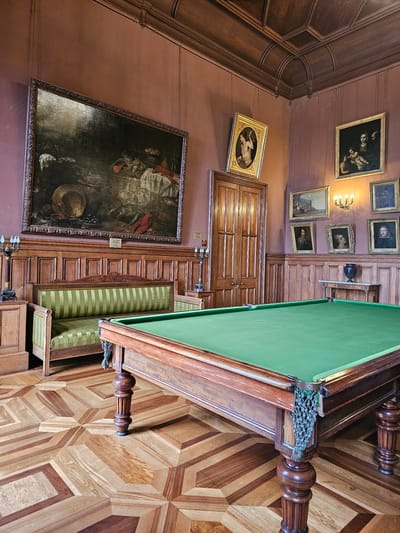 The image depicts an ornate room in what appears to be a historic building. The walls are reddish-brown, decorated with several framed paintings of various sizes. A large, dark painting is prominently displayed on the left side of the image. Other paintings hang along the upper wall.
A billiard table with a green felt surface occupies the right side of the image. Its wooden frame is intricately carved, and the table legs are elaborately turned. A net pocket is visible at the corner of the table. The floor is made of wooden parquet in a complex geometric pattern.
A wooden cabinet with drawers sits below the billiard table, and a wooden door with raised panels is visible in the background. Light fixtures are mounted on the walls.
The image likely represents a room in a palace or mansion, potentially located in or near Алупкинский городской совет, Ukraine, given the ornate architecture and historical artifacts. The scene is static and does not depict any people or immediate actions. The main focus is on showcasing the opulent interior design and the antique objects within the room.