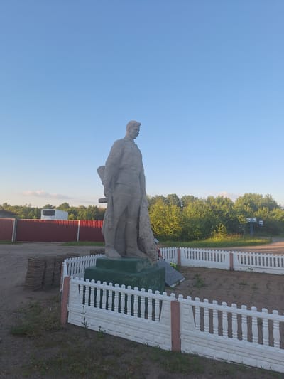 The image shows a statue of a man in military uniform, likely a war memorial, located in the city of Doktaravicki rural council, Belarus. The statue is made of stone and stands on a green pedestal, surrounded by a white decorative fence. Behind the statue, there's a red fence, a white refrigerator truck, and lush green trees under a clear blue sky. A bus stop sign is visible in the distance to the right. The area around the statue appears to be unpaved, with some stacks of concrete slabs to the left. The scene is bathed in natural daylight, suggesting it was taken during the day.
