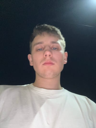 This is a close-up, low-angle, self-portrait photograph of a young Caucasian male taken at night in Seleevo, Russia. The subject is wearing a plain white t-shirt and his face is illuminated by an overhead light source, casting a slight glow on the dark background. The man has light brown hair, blue eyes, and fair skin. His facial expression is neutral, and he is looking directly at the camera. The image is slightly grainy due to the low light conditions. The background is uniformly dark, suggesting an outdoor setting at night, possibly a street or an open area within the city.