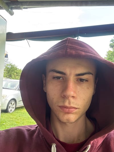 A young man with light skin, dark hair, and brown eyes is wearing a maroon hooded sweatshirt. He is positioned outdoors, possibly in Senoklas, Bulgaria, given the query context. The background includes green grass, a silver car, and parts of a building or structure, along with trees in the distance. The photo appears to be a self-portrait or taken at a close angle, with the subject's face prominent.