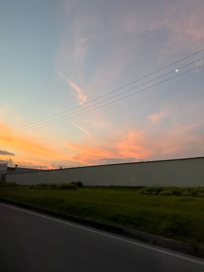 The image depicts an outdoor scene at dusk or dawn in Pitalito, Colombia, characterized by a vibrant sky and roadside elements. The upper half of the frame is dominated by a sky displaying a gradient of colors, transitioning from light blue on the left, through pale purple and pink, to strong orange hues on the right, indicative of a sunset or sunrise. Wispy, elongated clouds are present, reflecting these colors. Three parallel, dark power lines traverse the sky horizontally across the upper-middle section of the image. A distinct, bright circular light source, consistent with a moon or bright star, is visible in the upper right quadrant, positioned above the power lines.

Below the sky, a long, light-colored (likely concrete or stucco) wall extends horizontally across the mid-ground, acting as a clear boundary. To the left, behind the wall, a portion of a light-colored building with a flat roof is visible. Several dark, dome-shaped objects, possibly surveillance cameras or small satellite dishes, are mounted along the top edge of this building and intermittently along the top of the wall.

In front of the wall lies a strip of green grass, showing variations in shade and texture. Some darker, bush-like vegetation is visible at the base of the wall. The immediate foreground features a dark asphalt road surface, with a visible white line marking its edge and separating it from the grassy area. No people, discernible actions, or interactions are visible in the scene.