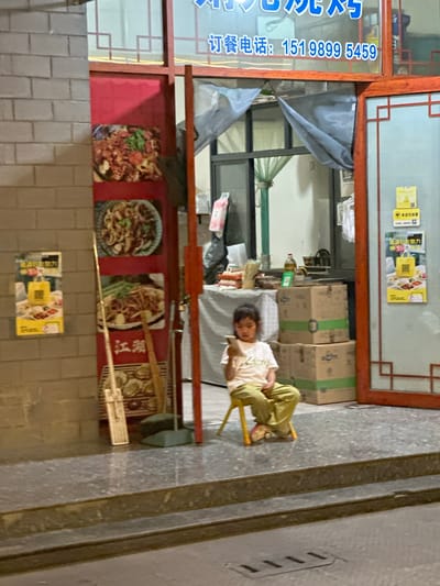 In Eshan Yi Autonomous County, China, a young girl sits on a small yellow stool, engrossed in her mobile phone. She wears a light-colored t-shirt and olive-green pants. The scene unfolds on a paved area in front of a shop with a red door frame and windows. Posters displaying food items are visible on the wall to the left of the entrance. Inside the shop, stacks of cardboard boxes are present, and a table is covered with a patterned cloth. A broom and dustpan are propped against the wall near the door. The overall environment appears to be an outdoor space adjacent to a commercial establishment.